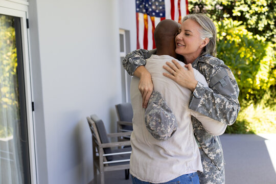 Diverse couple embracing on front porch, woman in camouflage holding patrol cap near American flag