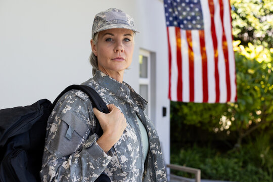 Standing on porch, female soldier in digital camo holding backpack near American flag, copy space