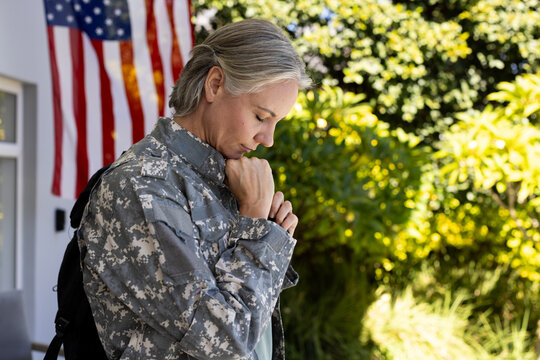 Mid-adult woman standing, bowing and clasping hands on porch by American flag wearing camo jacket