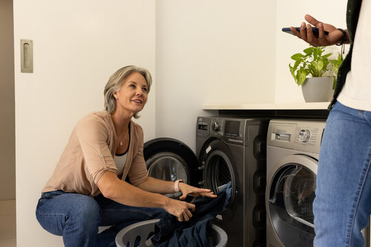 Couple loading laundry in home nook while woman crouching at washer and partner holding smartphone