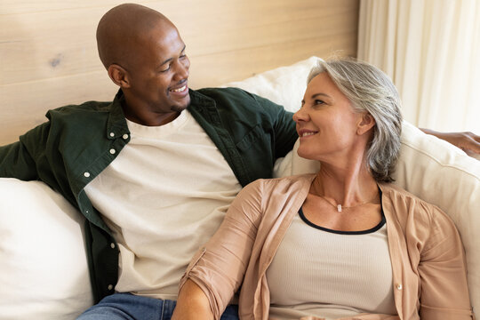 Diverse couple sitting on light sofa in living room, wearing casual clothes and pendant necklace