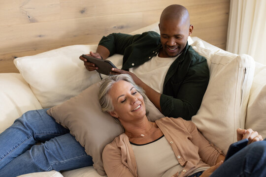 Diverse couple reclining on cream sofa at home, holding tablet with folio cover and smartphone