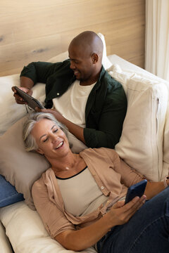 Diverse couple reclining on light-colored sofa in living room, using tablet and smartphone