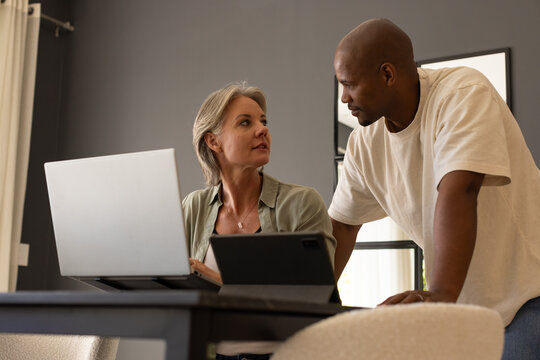 Diverse couple working and conversing at home dining table with silver laptop on stand and tablet