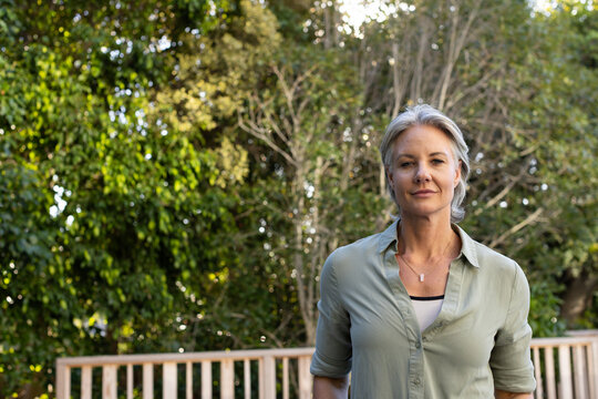 Senior woman standing on deck by railing facing camera, wearing sage shirt, pendant, copy space