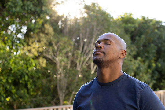 Man standing on wooden deck in backyard, wearing navy T-shirt, lifting chin to sunlight, copy space