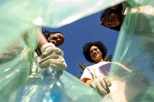 3 African women wearing gloves collecting plastic bottle near translucent green trash bag at park