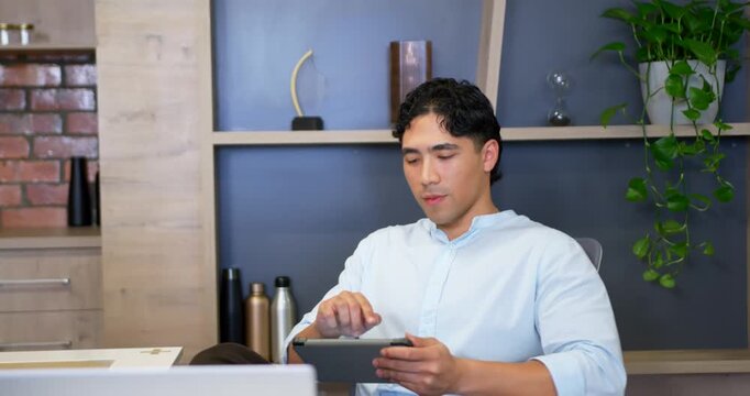 Adult Asian man sitting at desk checking tablet, tapping screen, leaning back smiling during break