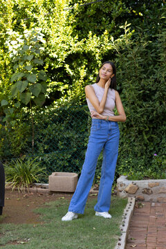 African American woman posing on backyard lawn in sleeveless top and jeans near planter block