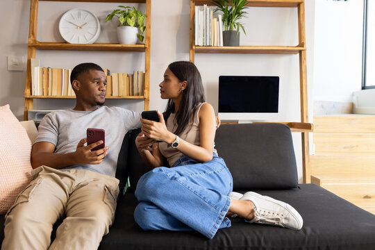 African American couple in casual clothes sitting on black sofa at home, holding smartphones