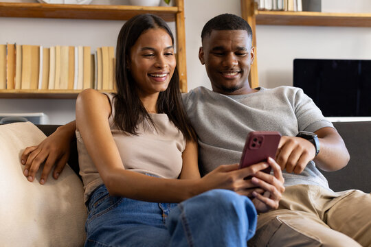 African American couple sitting on dark gray sofa in living room, holding pink phone and smiling