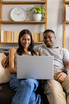 African American couple sitting on sofa at home using silver laptop with phone nearby, casual wear
