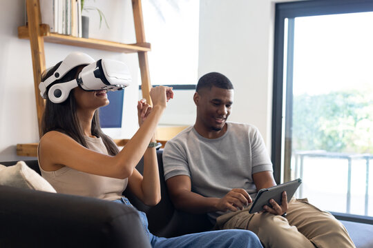 African American couple sitting on couch at home, woman with VR headset gesturing, man using tablet