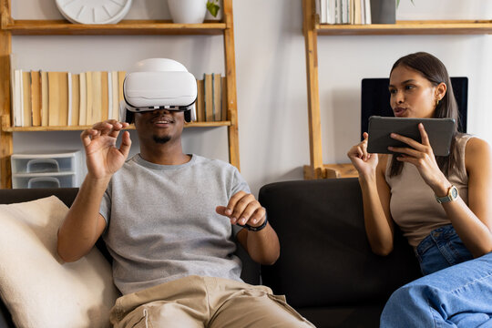African American couple sitting on dark sofa in living room using white VR headset and tablet