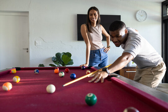 African American couple playing red-felt pool in living room, man lining up shot, copy space