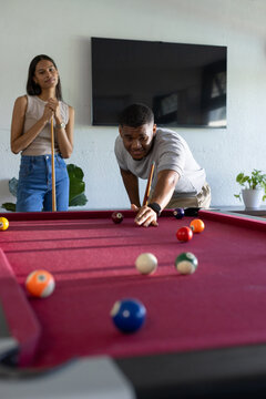 African American couple playing pool at home, man leaning over burgundy-felt table, two cues nearby
