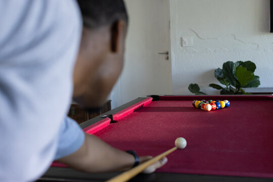 Adult African man leaning over red-felt pool table at home, aiming cue at cue-ball, copy space