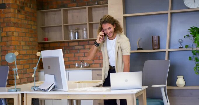 Man receiving call, holding phone, glancing at laptop and papers on desk in office, discussing work