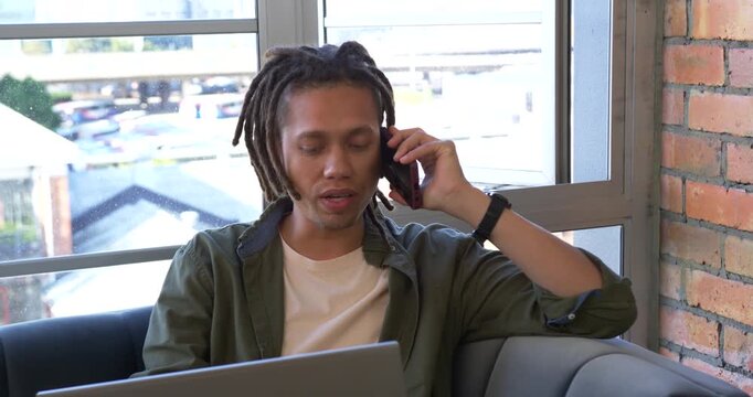 Adult African American man taking phone call for work, sitting on bench by window checking laptop