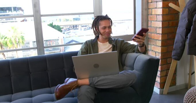 Man receiving call, sitting on tufted-sofa at window, speaking on red phone while working on laptop