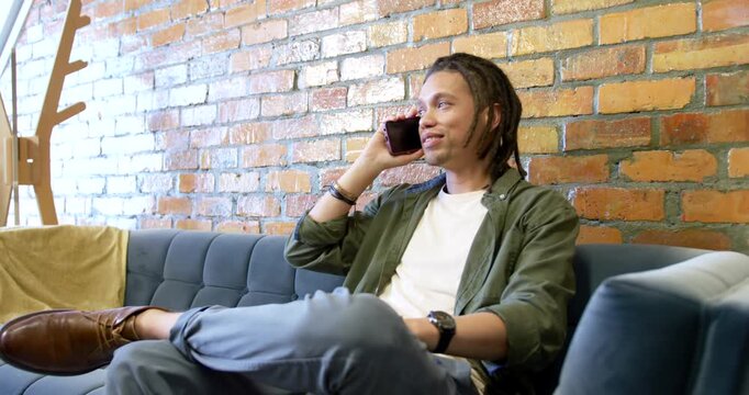African American man taking smartphone call, reacting to amusing comment and smiling on tufted sofa