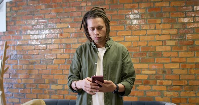 Man with dreadlocks in green shirt holding pink smartphone tapping, reading texts, smiling at home
