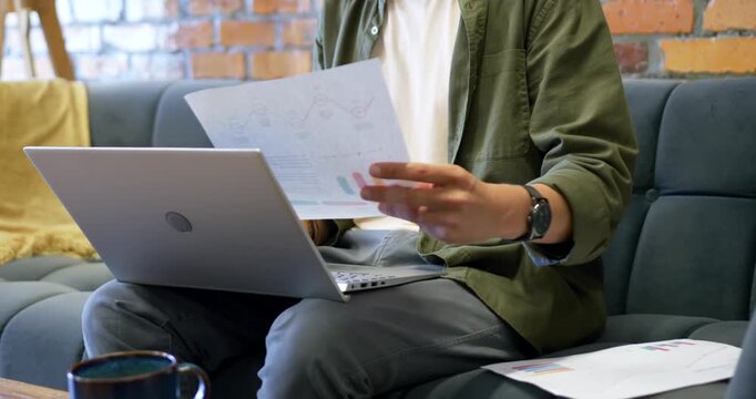 African American man holding printed charts and comparing with laptop on couch while analyzing data