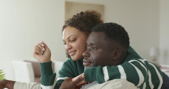 African American couple cuddling sofa spotting show, woman in green sweater holding remote watching