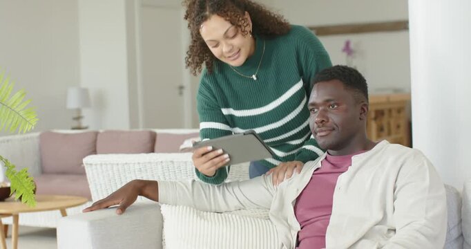 Diverse couple sitting on sofa, woman entering carrying tablet, guiding man while sharing photos