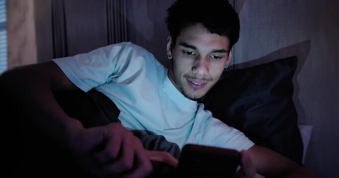African American man lying on bed with dark pillow, phone screen drawing glance, checking messages