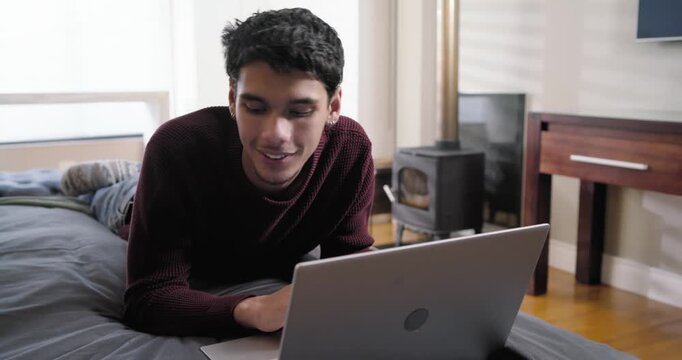 Man mid-20s in burgundy sweater lying on bed, typing on silver laptop holding card confirming order