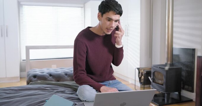 Man in his 20s taking smartphone call for work while typing on laptop gesturing on bed
