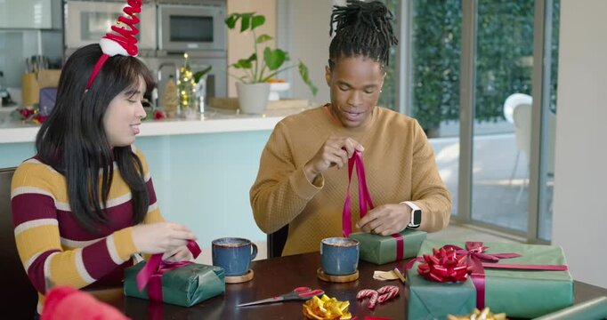Diverse couple wrapping green gifts at kitchen table, tying red ribbon while preparing for holiday