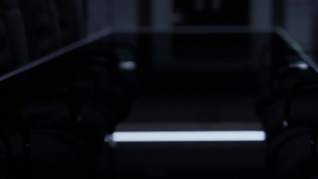 In a conference room, a glass table reflects dim light while chairs sit around it. The space lacks bright colors and the atmosphere feels serious.