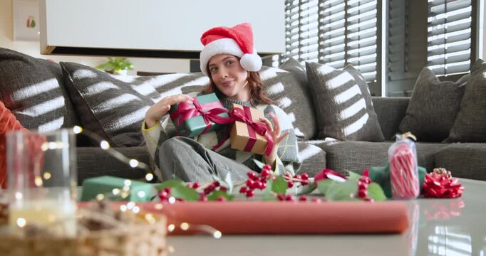Adult woman in Santa hat sitting on sofa holding two gifts, adjusting ribbons while wrapping gifts