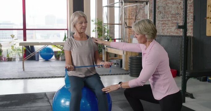 Mature female trainer guiding client doing band pulls on blue ball in rehab studio for posture