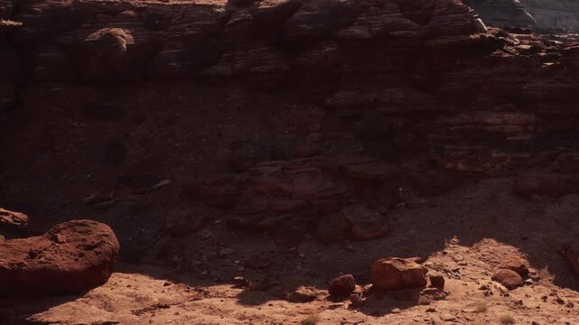 The scene shows a rocky area in a desert with various stones and soil. The sunlight creates shadows on the ground while the rocks stand tall. A clear sky can be seen above.