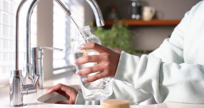 African American teen girl turning faucet, filling glass bottle, drinking water to hydrate at home