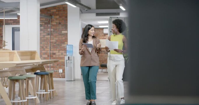 Diverse female coworkers walking as partition moving aside, reviewing tablet and papers in office