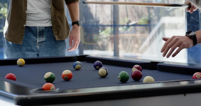 Two male friends watching right player breaking balls on pool table after aiming for friendly game