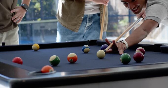Male friends cheering while right player lining up and striking cue ball to pocket numbered balls
