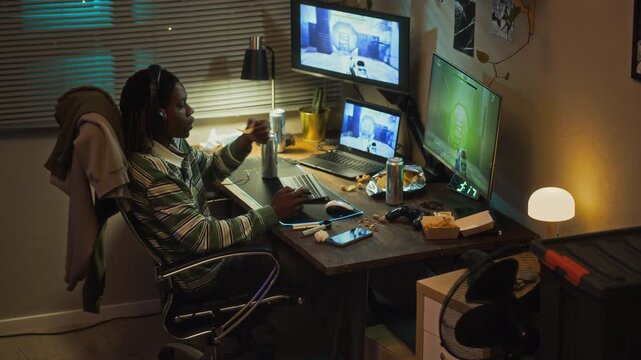African American teenage boy playing video games late at night at messy desk with multiple screens, eating snacks and drinking soda. Video suits projects on screen addiction and youth gaming habits