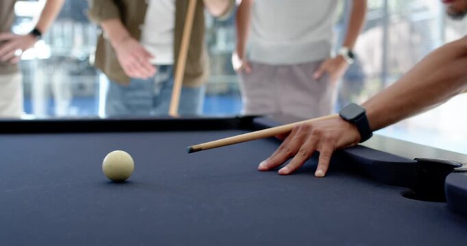 Diverse men watching shooter lining cue on pool table, striking cue ball to sink striped ball