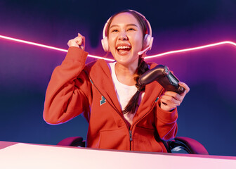 Host channel of smiling beautiful Asian girl streamer with joystick playing with raising fist up winning Esport skilled team players wearing headphones pastel media online in neon room. Stratagem. © InfiniteFlow