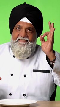 Indian Senior Male Sikh Chef in Traditional Attire Sitting at Table, Pointing at Empty Plate and Showing OK Sign on Green Screen