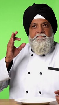 Indian Senior Male Sikh Chef in Traditional Attire Sitting at Table, Pointing at Empty Plate and Showing OK Sign on Green Screen