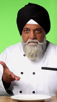 Indian Senior Male Sikh Chef in Traditional Attire Sitting at Table, Pointing at Empty Plate and Showing OK Sign on Green Screen