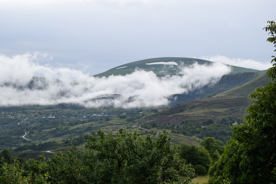 View of the mountainous landscape of Posof with low-hanging clouds, lush green slopes, and a small village nestled in the valley in Posof, Ardahan, Turkiye.