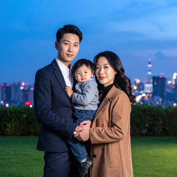 A stylish Asian family with a young toddler poses warmly at twilight against a vibrant city skyline. This elegant portrait captures modern love, success, and domestic harmony in urban life.