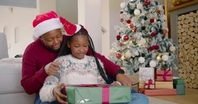 African American dad nudging gift into youth daughter's lap at home she inspecting ribbon smiling